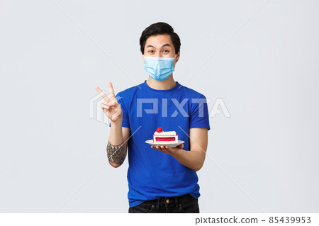 Different emotions, social distancing, self-quarantine on covid-19 and lifestyle concept. Handsome happy asian guy in medical mask eating takeaway at home, holding cake and show peace sign 85439953