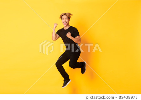 Full-length of happy attractive asian guy in black clothes jumping and celebrating victory, achieve goal, standing yellow background, triumphing 85439973