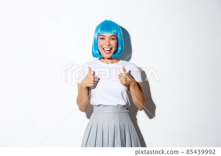Cheerful cute asian girl in blue wig looking satisfied, smiling and showing thumbs-up, wearing halloween outfit, standing over white background 85439992