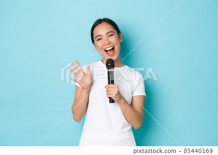 Lifestyle, people and leisure concept. Cheerful smiling asian girl enjoying karaoke night. Japanese female student singing song in microphone with happy emotions, standing light blue background 85440169