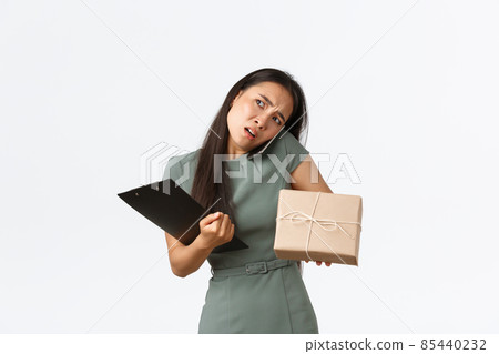 Busy confused asian woman answering phone call, talking with client puzzled, holding delivery box, prepare package for shipping, checking-in with customer, standing white background Busy confused asian woman answering phone call, talking with client puzzled, holding delivery box, prepare package for shipping, checking-in with customer, standing white background 85440232