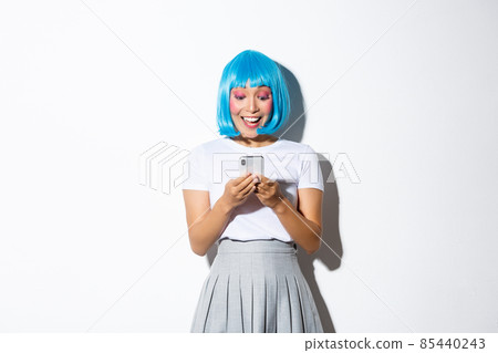 Image of attractive asian girl looking at mobile phone screen with happy and surprised face, standing in blue anime wig and halloween costume 85440243