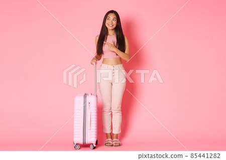 Travelling, holidays and vacation concept. Full-length of dreamy attractive asian girl explore country while being abroad, pointing finger left and smiling curious, standing with suitcase, pink wall 85441282