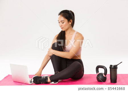 Portrait of slim healthy asian girl practice fitness at home, sitting on rubber mat with kettlebell and water bottle, follow online fitness coach instructions, using laptop to find yoga tutorials Portrait of slim healthy asian girl practice fitness at home, sitting on rubber mat with kettlebell and water bottle, follow online fitness coach instructions, using laptop to find yoga tutorials 85441668
