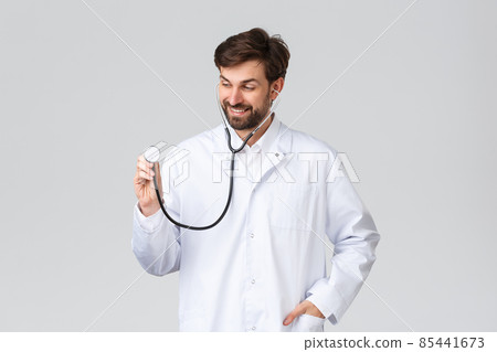 Hospital, healthcare workers, covid-19 treatment concept. Handsome doctor with beard in white scrubs, wearing stethoscope to listen patient lungs, treat clients in clinic, smiling, grey background 85441673