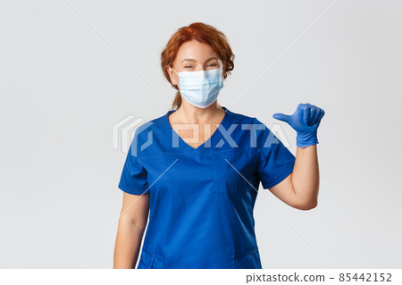 Medical workers, covid-19 pandemic, coronavirus concept. Smiling pleasant nurse, female professional doctor in medical mask, gloves and scrubs pointing at herself, being real pro, grey background 85442152