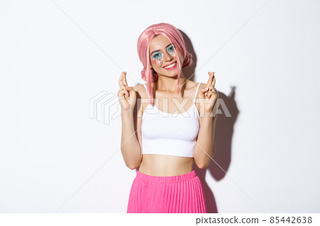 Portrait of lovely smiling woman in halloween costume, pink wig and bright makeup, looking hopeful at camera and making wish with fingers crossed 85442638