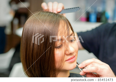 The hairdresser cuts the hair of a brunette woman. Hairstylist is cutting the hair of female client in a professional hair salon, close up. 85451473