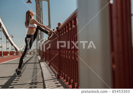 Tranquil slim woman stretches legs by handrail on emty footbridge Tranquil slim woman stretches legs by handrail on emty footbridge 85453303