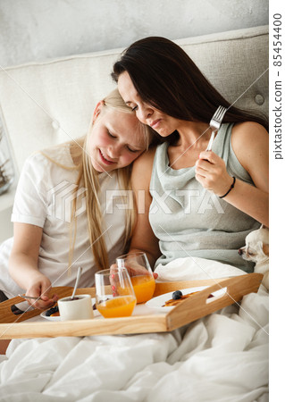 Having family breakfast in bed concept. Young happy mother eating pancakes and drinking orange juice with her teenage daughter. Woman and girl sitting in the bedroom in the morning. Having family breakfast in bed concept. Young happy mother eating pancakes and drinking orange juice with her teenage daughter. Woman and girl sitting in the bedroom in the morning. 85454400