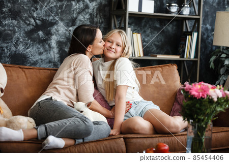 Mother and daughter relationship concept. A woman and a teenage girl are sitting in front of the TV on the couch and have fun talking. The family spends time at home. Little dog lying on laps Mother and daughter relationship concept. A woman and a teenage girl are sitting in front of the TV on the couch and have fun talking. The family spends time at home. Little dog lying on laps 85454406