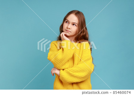 Portrait of adorable thoughtful little girl stands holds chin, looking away with pensive expression, wearing yellow casual style sweater. Indoor studio shot isolated on blue background. 85460074