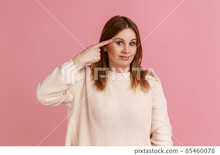 Portrait of unhappy blond woman touching finger near temple feeling herself stupid, dissatisfied showing dumb gesture, wearing white sweater. Indoor studio shot isolated on pink background. 85460078