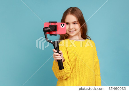 Portrait of little girl with positive expression, holding steadicam and smart phone, streaming, wearing yellow casual style sweater. Indoor studio shot isolated on blue background. 85460083