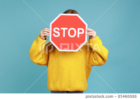 Portrait of unknown little kid covers face with Stop symbol, anonymous child holds red traffic sign, wearing yellow casual style sweater. Indoor studio shot isolated on blue background. Portrait of unknown little kid covers face with Stop symbol, anonymous child holds red traffic sign, wearing yellow casual style sweater. Indoor studio shot isolated on blue background. 85460088