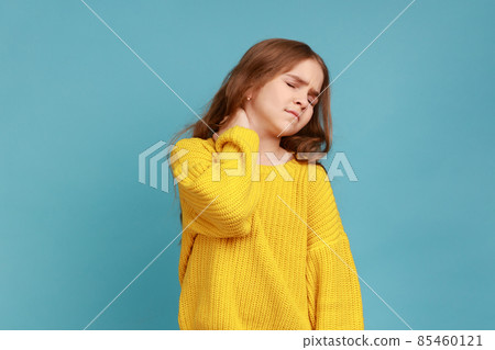 Portrait of little girl massaging painful back, feel discomfort in neck, child sedentary lifestyle, wearing yellow casual style sweater. Indoor studio shot isolated on blue background. Portrait of little girl massaging painful back, feel discomfort in neck, child sedentary lifestyle, wearing yellow casual style sweater. Indoor studio shot isolated on blue background. 85460121
