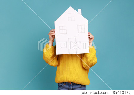 Portrait of unknown little kid hiding behind paper house, affordable housing program, advertisement, wearing yellow casual style sweater. Indoor studio shot isolated on blue background. Portrait of unknown little kid hiding behind paper house, affordable housing program, advertisement, wearing yellow casual style sweater. Indoor studio shot isolated on blue background. 85460122