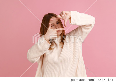Portrait of blond woman imitating to take picture, looking through finger frame with attentive view, focusing and cropping image, wearing white sweater. Indoor studio shot isolated on pink background. Portrait of blond woman imitating to take picture, looking through finger frame with attentive view, focusing and cropping image, wearing white sweater. Indoor studio shot isolated on pink background. 85460123