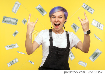 Oh my god, wow. Amazed woman with violet short hair showing rock and roll gesture looking at camera screaming, absolutely shocked of money rain falling from up. yellow background, studio shot 85460357