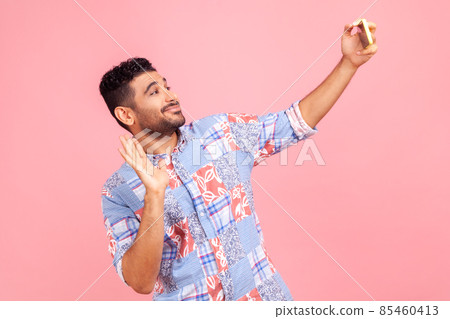 Handsome man with beard in blue casual style shirt talking on video call and waving hello gesture, having online conversation on mobile phone. Indoor studio shot isolated on pink background. Handsome man with beard in blue casual style shirt talking on video call and waving hello gesture, having online conversation on mobile phone. Indoor studio shot isolated on pink background. 85460413