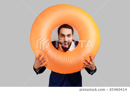 Bearded man holding rubber ring over her neck and looking at camera with surprised expression and opened mouth, wearing official style suit. Indoor studio shot isolated on gray background. Bearded man holding rubber ring over her neck and looking at camera with surprised expression and opened mouth, wearing official style suit. Indoor studio shot isolated on gray background. 85460414