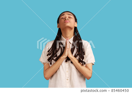Woman with black dreadlocks holding hands in prayer and pleading, looking up with eyes full of hope, asking apology, wearing white shirt. Indoor studio shot isolated on blue background. 85460450