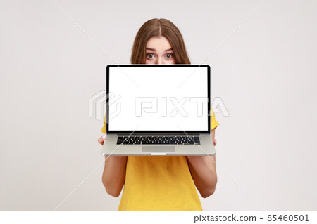 Positive unknown female in yellow T-shirt hiding half of face behind laptop with white empty display, looking at camera with big surprised eyes. Indoor studio shot isolated on gray background. Positive unknown female in yellow T-shirt hiding half of face behind laptop with white empty display, looking at camera with big surprised eyes. Indoor studio shot isolated on gray background. 85460501
