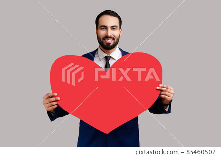 Portrait of smiling handsome bearded man holding big red heart, expressing positive romantic emotions, wearing official style suit. Indoor studio shot isolated on gray background. Portrait of smiling handsome bearded man holding big red heart, expressing positive romantic emotions, wearing official style suit. Indoor studio shot isolated on gray background. 85460502