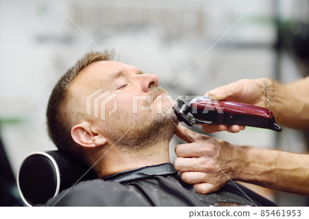Barber master is shaving of handsome mature bearded man in salon. Hair artist making hairstyle for person in barbershop. Services of professional stylist. Barber master is shaving of handsome mature bearded man in salon. Hair artist making hairstyle for person in barbershop. Services of professional stylist. 85461953