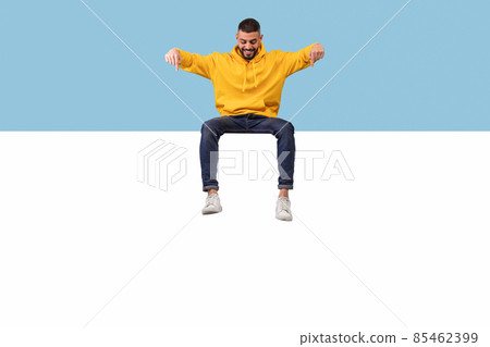 Happy middle eastern man sitting on big blank advertising board, pointing and looking down at empty space Happy middle eastern man sitting on big blank advertising board, pointing and looking down at empty space 85462399