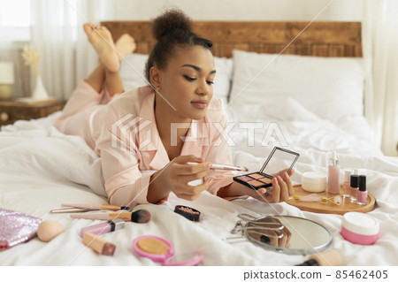 Daily morning routine. Pretty african american woman in pajamas applying makeup, holding brush and eyeshadow palette Daily morning routine. Pretty african american woman in pajamas applying makeup, holding brush and eyeshadow palette 85462405