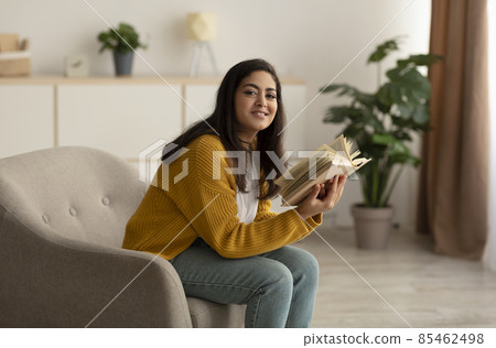Peaceful stay home pastimes. Happy arab lady reading book, sitting on armchair in living room and smiling at camera Peaceful stay home pastimes. Happy arab lady reading book, sitting on armchair in living room and smiling at camera 85462498