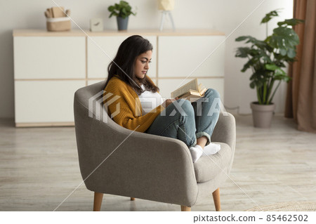 Focused arab lady sitting in armchair, reading book in living room, enjoying lazy morning with interesting story Focused arab lady sitting in armchair, reading book in living room, enjoying lazy morning with interesting story 85462502