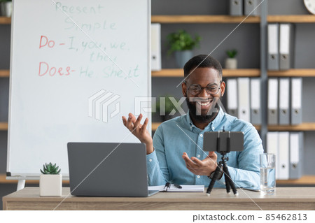 Smiling millennial black male teacher in glasses look at laptop, phone webcam in home office interior Smiling millennial black male teacher in glasses look at laptop, phone webcam in home office interior 85462813