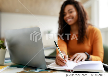 Black woman sitting at desk, using pc writing in notebook Black woman sitting at desk, using pc writing in notebook 85462818