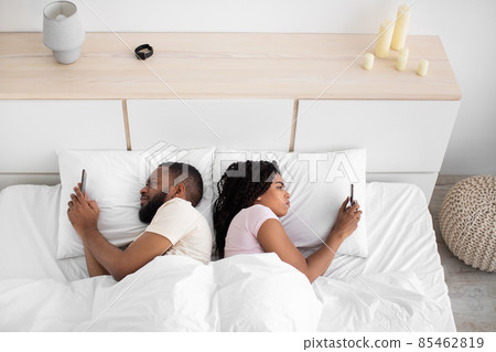 Sad young black guy and lady lie back to back, looks at smartphones on bed in bedroom interior Sad young black guy and lady lie back to back, looks at smartphones on bed in bedroom interior 85462819
