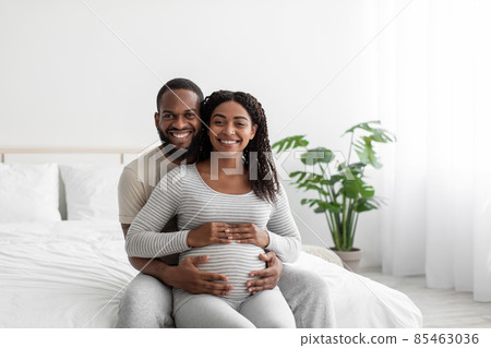 Satisfied smiling handsome millennial african american guy hugs pregnant lady with belly, sit on bed Satisfied smiling handsome millennial african american guy hugs pregnant lady with belly, sit on bed 85463036