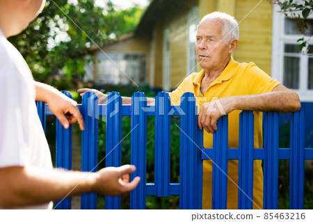 Neighbor conversation. Two smiling men breezily chatting near fence of rural house Neighbor conversation. Two smiling men breezily chatting near fence of rural house 85463216