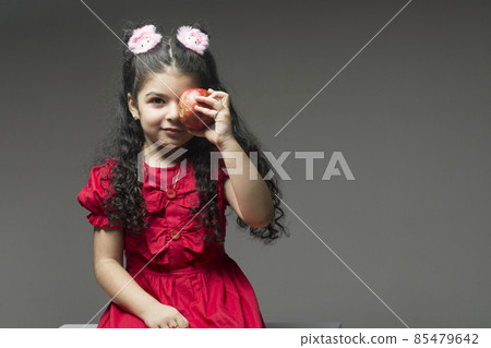 Little girl with red dress holding a pomegranate in front of her left eye Little girl with red dress holding a pomegranate in front of her left eye 85479642