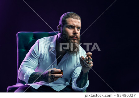 Portrait of thinking bearded man who smoke cigar holds glass of whiskey while sittingin armchair. Portrait of thinking bearded man who smoke cigar holds glass of whiskey while sittingin armchair. 85480327