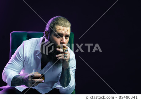 Portrait of handsome bearded man who smoke cigar holds glass of whiskey while sittingin armchair. Portrait of handsome bearded man who smoke cigar holds glass of whiskey while sittingin armchair. 85480341