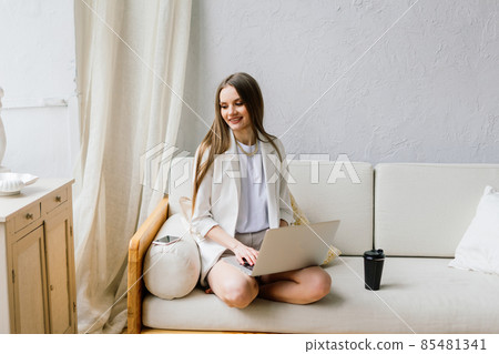 Beautiful young woman working on laptop computer while sitting at the living room, drinking coffee Beautiful young woman working on laptop computer while sitting at the living room, drinking coffee 85481341