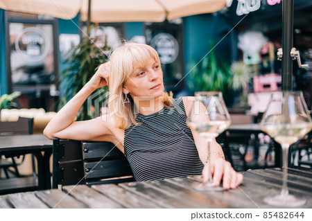 Thoughtful adult mature woman sitting in bar outdoors with wine glasses and blurry restaurant background scene, drinking white wine. Summer sunny day on patio. People lifestyle. Thoughtful adult mature woman sitting in bar outdoors with wine glasses and blurry restaurant background scene, drinking white wine. Summer sunny day on patio. People lifestyle. 85482644