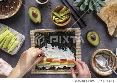 Cooking Sushi rolls at home with salmon, avocado and cucumber. Female hand preparing avocado sushi, step by step. 85485483