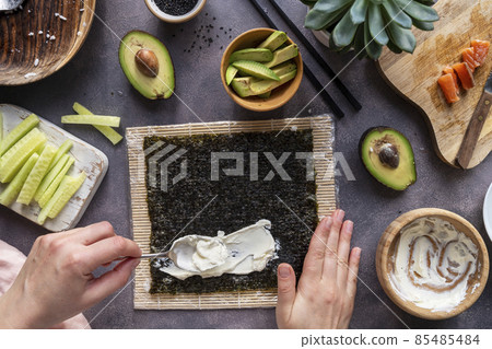 Cooking Sushi rolls at home with salmon, avocado and cucumber. Female hand preparing avocado sushi, step by step. 85485484