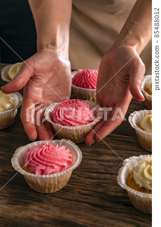Female hands keeps vanilla cupcake with pink cream. Just cooked cakes on wooden table. Vertical frame. 85488012