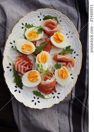 Italian salad. Boiled eggs with smoked ham and arugula on white plate. Dark wooden background 85489951