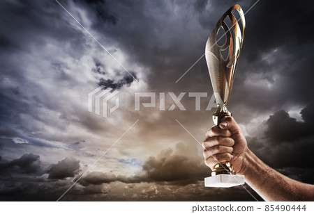 Man's hand is holding gold cup against of the gloomy stormy sky. 85490444