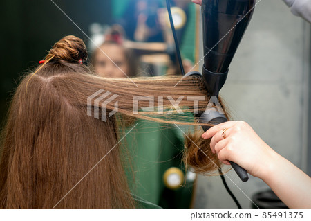A hairdresser is drying long brown hair with a hairdryer and round brush in a hairdressing salon. 85491375