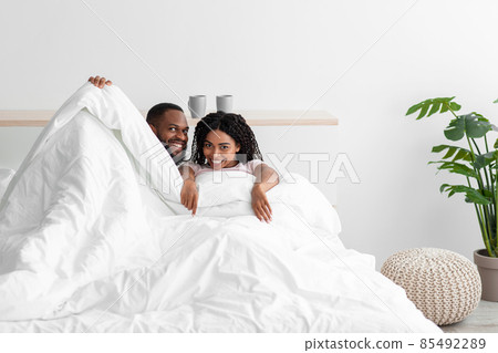 Glad smiling young black man and woman covered with blanket on bed enjoy happy moment at weekend 85492289